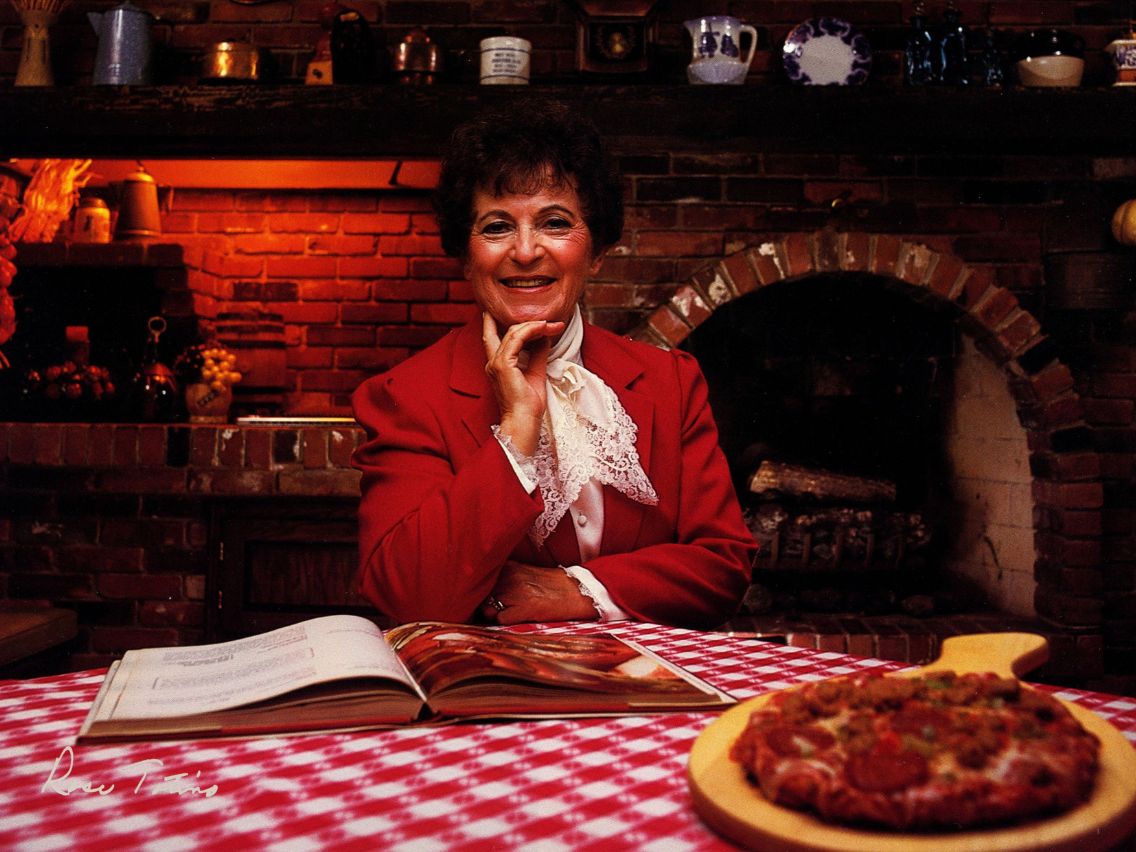 Rose Totino with her hand on her chin smiling, sitting at a table with a red and white checkered tablecloth with an open recipe book and cooked pizza sitting in front of her. In the background you can see the dimly lit brick pizza oven.